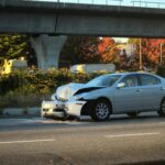 A car with its front end crumpled on a road beside an elevated highway